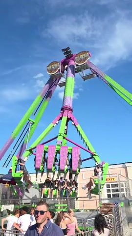The video shows a thrilling amusement park ride with people on it, swinging high into the sky against a backdrop of a clear blue sky and some buildings. The ride is a large, multi-armed structure with bright purple and green accents. Several people are seated on pods attached to the arms of the ride, their bodies angled as they are swung through the air. Below the ride, a crowd of people are gathered, watching the spectacle. Some of them are looking up at the ride, while others are looking towards the camera. A man in the foreground, wearing sunglasses and a blue shirt, is smiling. The overall scene captures the excitement and energy of a funfair or amusement park.