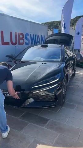 A man in jeans and a navy t-shirt is working on the front license plate of a sleek black car, which is parked on a stone tile surface. The car's back hatch is open. The car is labeled "DENZA Z9 GT". Behind the car is a white banner with the text "CLUB SWAN" in large red and blue letters.  Two white vertical flags are also in the background. There is a small hill covered in greenery in the background. The sky is blue with scattered white clouds. The man is crouched down and appears to be adjusting the license plate.