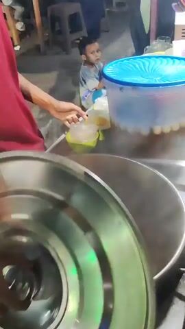 A medium shot shows a person in a red shirt holding a scoop over a large pot filled with steaming liquid. The person is in the process of transferring something from a small green bowl into the pot. In the background, a child is visible, looking towards the person with the scoop. To the right of the pot is a large blue-lidded container. The environment appears to be an outdoor food stall with dim lighting.