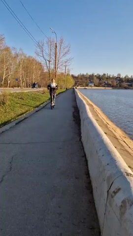 A person rides an electric scooter on a paved path alongside a body of water. The path is bordered by a low concrete wall on the water side and a grassy area with trees on the other. The water is calm with small ripples, and the opposite shore is visible in the distance, featuring trees and buildings. The sky is clear and blue. A car drives by on a road adjacent to the path. The camera pans right following the line of the water as the scooter moves past.
