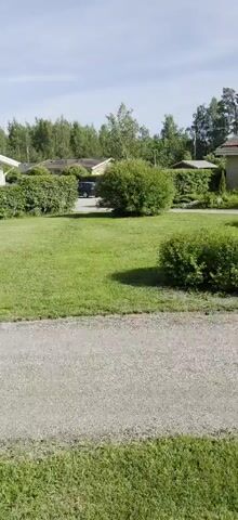 This is a panning video of a suburban or rural Finnish neighborhood on a sunny day. The scene features several single-story houses with light-colored facades and dark roofs, surrounded by lush green lawns and neatly trimmed hedges. A gravel driveway runs along the bottom of the frame, with a well-maintained lawn on the other side.

In the foreground, the camera pans across a green lawn and a gravel path. In the middle ground, several houses are visible, with their garages and hedges. A dark-colored car is parked in one of the driveways. A tall flagpole with a flag flying is visible on the left side of the frame. A lamppost is also visible in the distance.

The background is dominated by a dense line of trees, suggesting a wooded area bordering the residential properties. The sky is a clear blue with a few wispy clouds, indicating fair weather. The time of day appears to be late morning or early afternoon, given the bright sunlight and the angle of the shadows.

The overall impression is one of peaceful domesticity and well-kept surroundings. There is no text visible in the video. The location is identified as Uusikaupunki, Finland, which aligns with the architectural style of the houses and the prevalence of trees and greenery.