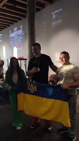 In a dimly lit room, three individuals are posing for a photograph, holding a Ukrainian flag. A woman on the left, wearing a green tracksuit, smiles broadly. A man in the center, wearing a black t-shirt and red pants, displays a medal around his neck and his left fist is raised. To his right, another man in a beige t-shirt, with a muscular physique, also raises his left fist. The Ukrainian flag, with its blue and yellow colors and the Tryzub emblem, is held prominently by the two men. In the background, a large screen displays text and images, possibly related to an event called "BABAEV'S CUP". The floor is made of dark wood.