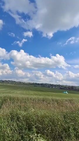 The video shows a wide outdoor landscape under a bright blue sky filled with fluffy white clouds. In the foreground, there is a field of tall, dry grass and some green vegetation. In the mid-ground, there is a large, vibrant green field, likely a pasture or meadow. Further in the distance, several colorful kites are visible on the green field. There appear to be two kites, one light blue and one orange, set up in the field. The background shows a gently rolling landscape with some scattered houses or buildings along the horizon. The overall scene is one of a pleasant, possibly windy day outdoors.