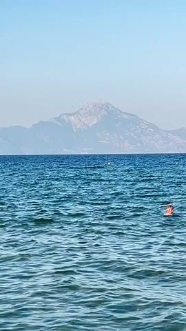 The video shows a wide shot of a clear blue ocean on a sunny day. The water is calm with small ripples. In the foreground, the sandy bottom of the ocean is visible through the clear water. In the middle ground, several people are swimming and wading in the ocean. Some are wearing orange inflatable flotation devices. In the background, a large, hazy mountain range with snow-capped peaks is visible across the water under a clear blue sky. The camera appears to be static, facing the ocean and the mountain range.
