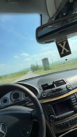 The video captures a first-person perspective from the driver's seat of a Mercedes-Benz car, showing the dashboard, steering wheel, and a clear view through the windshield. An "Areon X" air freshener hangs from the rearview mirror. The car is driving on a paved, single-lane road that stretches straight into the distance, flanked by green fields and trees. In the background, there are rolling hills under a bright blue sky with scattered white clouds. To the right of the road, a tall, wooden structure, possibly a hunting stand or observation tower, is visible among the trees. The sun is shining brightly, indicating daytime. The scene suggests a drive through a rural landscape in Kovachite, Bulgaria.