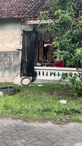 The video shows an exterior view of a house, with a porch area and surrounding greenery. In the background, a person wearing a striped shirt and red pants appears to be moving around the porch. Various objects are visible, including plants, a broom, a hose, and what appears to be a trash can. The house has a tiled roof and textured walls. The ground is paved with stones and covered in some grass and debris. A utility pole with markings is also visible on the right side of the frame.