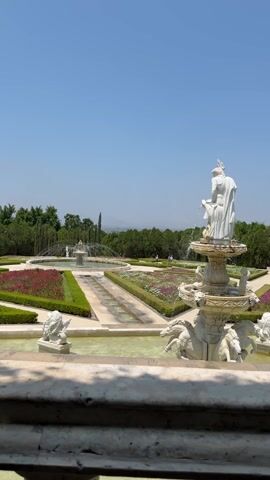 The video shows a picturesque view of a European-style garden on a sunny day. In the foreground, a white balustrade partially obscures the view, offering a glimpse of the garden through its columns. A statue, partially cut off by the frame, stands on a fountain to the right. Below it are additional marble decorations and smaller fountains.  

The garden itself is characterized by geometric shapes of flower beds filled with red and pink flowers, interspersed with rectangular hedges. Water features like small pools and fountains create symmetry within the layout.  

In the background, lush green trees and distant mountains are visible against a clear blue sky. A woman wearing pink walks briefly on the upper left side of the frame. The overall scene evokes a sense of tranquility and grandeur.