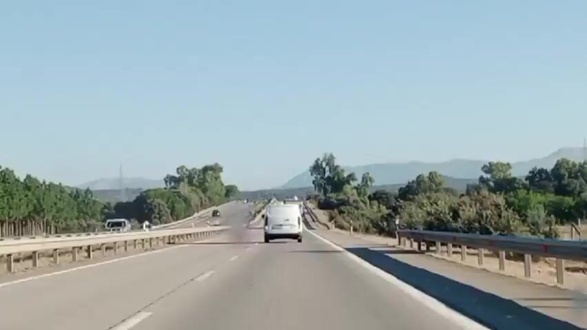 A first-person perspective shot shows a white van driving down a highway on a clear, sunny day. The van is in the center of the frame, driving away from the camera. The highway has multiple lanes and is surrounded by trees and hills in the distance. Other vehicles, including cars and trucks, can be seen on the highway.
