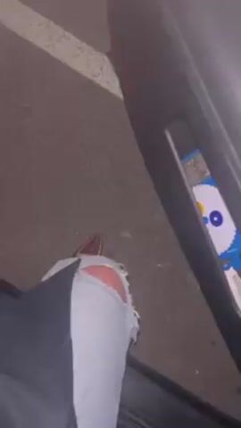 A person with ripped jeans and brown pointed-toe shoes exits a grey car in a parking space. The person steps out of the car onto the asphalt and stands next to the open car door. The parking space is marked with a white line.