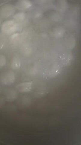A close-up shot shows a pot of boiling water with numerous small, white dumplings. The dumplings are submerged in the water and appear to be cooking, indicated by the bubbles rising to the surface. The lighting is dim, and the water is cloudy, obscuring the bottom of the pot and the dumplings at the very bottom. The dumplings are arranged in multiple layers, filling most of the visible area of the pot. The texture of the dumplings is smooth, and their shape is consistent, with a slightly pointed top. The boiling action creates a dynamic and somewhat chaotic visual effect in the water, with ripples and splashes visible.
