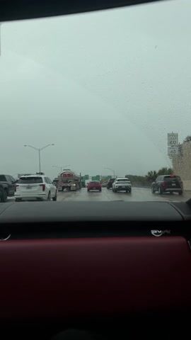 From the interior of a vehicle, the video shows a cloudy day on a highway. The windshield is covered in raindrops, and the wipers move. Traffic is visible ahead, including cars, SUVs, and a tanker truck with "SSI Chemicals" printed on its side. Streetlights line the side of the road, and green highway signs are overhead. The dashboard of the vehicle is in the foreground, with a red leather trim visible. An "SVR" badge is also visible on the dashboard.