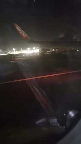 The video was shot from the window of an airplane. It shows the plane on a dark runway, the lights of an airport in the background, and the wing of another EasyJet airplane visible through the window. The colors of the runway lights appear shifted toward red due to the cabin lights. The window appears smudged.