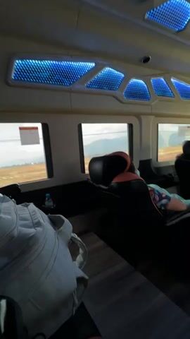 The video shows the interior of a bus or van, viewed from the back. The interior features a modern design with blue LED lighting in a patterned ceiling.  There are rows of black leather seats, some of which are occupied by passengers who appear to be relaxing.  A white backpack sits on the floor in the foreground.  The camera pans upward and backward.  Windows along the sides show glimpses of an outdoor landscape with brown fields and distant hills.  The vehicle seems to be in motion, based on the changing scenery outside the windows. A driver is visible through the front windshield.