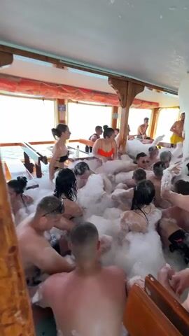 A lively foam party is taking place on the interior deck of a boat in Menderes, Turkey. Numerous young adults, both male and female, are immersed in a thick layer of white foam that fills much of the space. Most individuals are in swimwear, such as bikinis and swim shorts.

A foam cannon, visible centrally and toward the back, continuously dispenses more foam into the party area. People are seen smiling, laughing, and interacting with each other and the foam, suggesting a joyous and energetic atmosphere. The boat features wooden paneling and large windows, through which the bright blue sea and sky are visible, indicating it's daytime. In the background, a bar area is discernable, with more people and drinks. The scene depicts a fun, celebratory event on the water. No visible text is present.