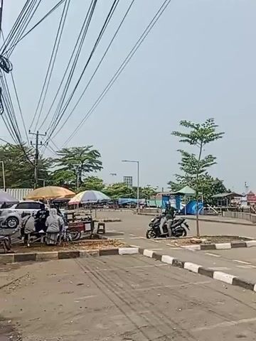 The video shows a street scene with a body of water and boats in the background. There are several parked motorcycles and some street vendors selling food from carts under umbrellas. A person is seen riding a motorcycle down the street. Power lines are visible overhead. The sky is clear and the weather appears to be sunny. The overall environment suggests a coastal town or market area.
