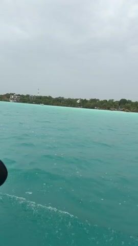 This is a POV video from a boat moving through turquoise water. On the horizon, there is a tree-lined shore with scattered buildings. The sky is overcast.