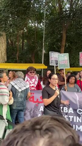 The video shows a crowd of people gathered outdoors, likely at a demonstration or rally. The focus is on a woman who is speaking into a microphone. She is wearing glasses and a black shirt. Behind her is a banner with Arabic writing and images of people. Several people behind the woman are holding signs. One sign reads "20 ANOS DOS CRIMES DE MAIO" and another reads "Absolvição de Zé Maria, já!". People in the crowd are wearing red and white scarves, some with the acronym CBR. The setting is a park or tree-lined area. The camera appears to be held by someone in the crowd.