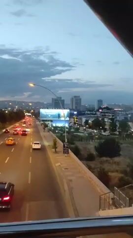 The video shows a view from above looking down at a multi-lane highway at dusk. Cars are driving in both directions, their headlights and taillights illuminated. Tall modern buildings are visible on the right side of the highway, along with streetlights and some trees. The sky is cloudy and the overall lighting suggests it is either early morning or late evening. A large billboard is visible on the right side of the highway.