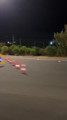The video shows a car driving on a paved surface at night, indicated by the dark sky and streetlights. There are several orange and white traffic cones set up, suggesting a driving course or a practice area. In the background, there are trees, utility poles, and lights from what appears to be a distant town or city. Several people in reflective vests are visible, likely supervising or participating in the activity. A white vehicle is also present. The car in the foreground is moving, and the camera appears to be inside another vehicle, capturing the scene from a side window. The car ahead performs a maneuver, possibly drifting or turning sharply around the cones. The overall impression is a controlled driving event taking place in an open area at night.