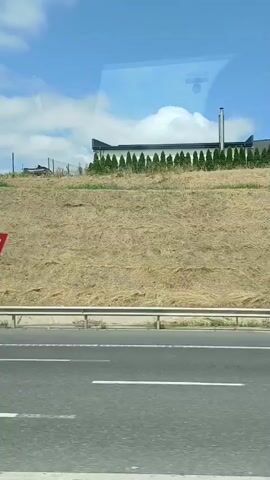 The video shows a view from a moving vehicle, likely a car or bus, looking out the side window. The scene is a roadside environment with a clear blue sky dotted with fluffy white clouds. In the foreground, a section of asphalt road is visible, marked with white directional arrows indicating movement. A metal guardrail separates the road from a steep, grassy embankment. The embankment is covered in dry, light-brown grass and sparse green bushes and trees. At the top of the embankment, several buildings and structures are visible, including what appears to be a building with a dark roof and white decorative elements along its facade. Further back on the hill, more greenery and glimpses of structures can be seen. The overall impression is of a sunny day in a rural or suburban area, possibly along a highway.