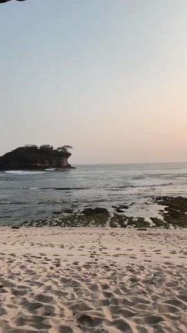 The video shows a serene beach scene at what appears to be sunset or sunrise. In the foreground, there is a sandy beach with some footprints visible. The sand is light brown in color. Beyond the beach, the ocean stretches out to the horizon. The water is calm with gentle waves lapping at the shore. There are exposed rocks and patches of green algae in the shallow water near the beach. In the distance, a small, rocky island with trees on top is visible. The sky is a pale, hazy color, suggesting the sun is low on the horizon. There are a few dark shapes in the water near the island that could be people or small boats. The overall atmosphere is peaceful and tranquil.