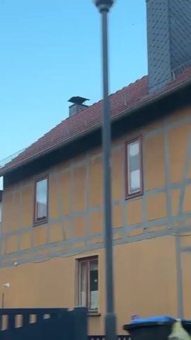The video shows the exterior of several buildings with a variety of architectural styles. The first building has a yellow facade with dark beams forming a half-timbered pattern. The second building has a dark grey, shingled exterior with white-framed windows and a large green bush in front of it. The third view shows a narrow gap between two buildings, one with a white facade and dark beams and the other with a dark grey shingled exterior. The final shot is of a long, red-tiled roof with a half-timbered facade below, against a light blue sky with a few wispy clouds. Two red brick chimneys are visible on the roof, and a gutter runs along the edge. The video appears to be taken from a moving vehicle, as evidenced by the slight blur and the changing perspective.