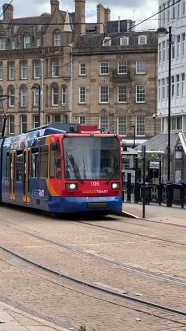 The video shows a blue, red, and yellow Supertram tram car pulling into the Cathedral station. The tram car is labeled “106” on the front. The stop is marked with signs that read "Cathedral." The tram travels on rail tracks embedded in a brick-paved street. In the background are several buildings with multiple stories and many windows.  A few people are visible at the station. The sky is overcast.