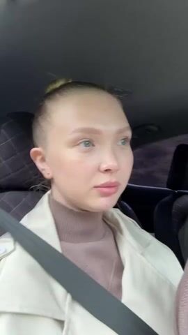 A young woman with light skin, blue eyes, and blonde hair pulled back in a bun is sitting in the passenger seat of a car. She is wearing a beige trench coat over a brown turtleneck. The car's interior is visible, including the quilted headrest of the seat and a grey seatbelt crossing her chest. The background outside the car appears to be a natural landscape with trees and possibly hills, suggesting a rural or semi-rural setting. The lighting is natural, indicating daytime, and the overall mood seems calm and contemplative. The woman is looking slightly off-camera to her right, with a neutral or slightly thoughtful expression. There is no visible text in the media file.