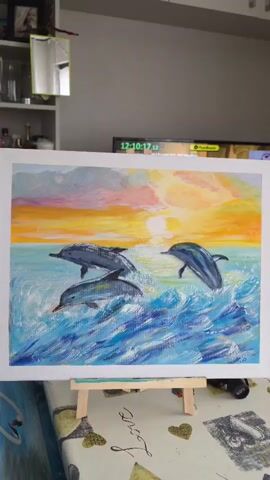 The video shows a painting of three dolphins jumping out of the water during a sunset. The painting is on an easel in front of a blurry background that appears to be a room with shelves and a television. The dolphins are depicted in shades of gray and blue, with white highlights on their bodies and in the splashing waves. The sunset is rendered in warm colors of orange, yellow, pink, and purple, with a bright sun visible on the horizon. The water is painted with vibrant blues and white, creating a sense of movement and energy.