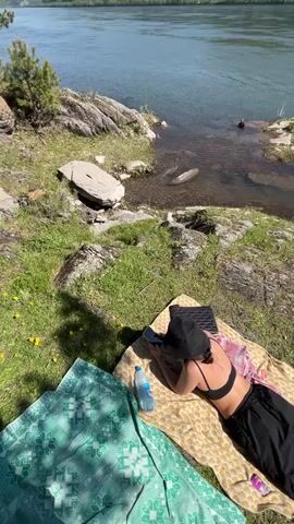 The video captures a sunny daytime scene on the grassy bank of a wide river in Слизнево, Russia.

A person is shown lying face down on a beige-patterned blanket spread on the grass. They are wearing a black sports bra or bikini top, black pants, and a black bucket hat. A blue water bottle and a smartphone are placed on the blanket next to them. A second green-patterned blanket is laid out nearby. The river, with its calm, clear water, flows beside the bank, which is characterized by green grass, rocks, and some sparse vegetation. The camera briefly shows the person's feet, clad in athletic shoes, before panning.

Further along the riverbank, another person is sitting in a folding camping chair, facing the water. Next to them, a small, portable grill is set on the ground, surrounded by several bags and other picnic or camping supplies. The expansive river stretches into the distance, with green, tree-covered hills and mountains visible under a clear blue sky.

The scene depicts a peaceful and relaxed outdoor activity by the river, possibly picnicking or simply enjoying the natural environment. No visible text is discernible on any objects.