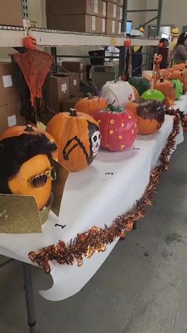 The scene is a table filled with decorated pumpkins in a warehouse-like setting. The table is covered with a white tablecloth and decorated with orange tinsel. There are at least ten pumpkins, each uniquely decorated, including one resembling Elvis Presley and another painted like a strawberry. The pumpkins are numbered in order from one to three. Behind the table are shelves stacked with boxes. In the background, there are people walking and standing.  The lighting is bright and fluorescent.
