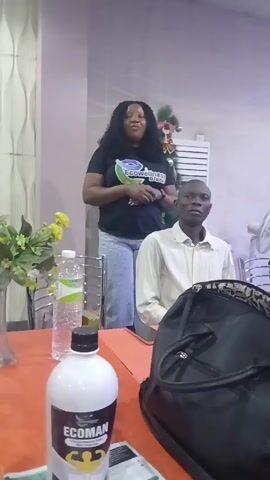 A woman wearing a black t-shirt and jeans is standing behind a table and speaking to a man sitting at the table. The woman's t-shirt has the logo "Ecowellness Global" on it. There is a bottle labeled "Ecoman" on the table, along with other bottles of water. There are flowers in a vase on the table. The man is wearing a white shirt and is sitting upright with his hands clasped in his lap.  Behind the woman is a Christmas tree. A black bag is on the table to the right. The table is covered with an orange tablecloth. The room appears to be indoors, with white walls and standard office-like fixtures.