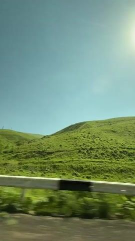 This is a horizontal video taken from a moving vehicle, showing a view of green rolling hills under a clear blue sky. The sun is visible in the upper right corner, causing a lens flare. A white and reflective guardrail runs along the bottom of the frame, with blurred greenery behind it. The hills are covered in lush green grass and appear to be uninhabited, with no structures or people visible. The motion of the vehicle creates a slight blur in the foreground and midground, emphasizing the sense of travel. The overall impression is one of a serene and vast natural landscape.
