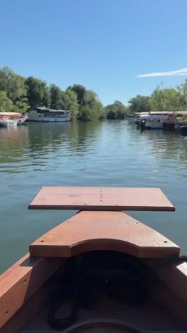 The video captures a first-person perspective from a small wooden boat moving slowly on a body of water. The front of the boat, featuring a small wooden table-like structure, is visible in the foreground. The water ahead is calm and reflects the clear blue sky. On both sides of the waterway, several larger boats are docked along the banks, which are lined with lush green trees and foliage. The sky is bright and clear with no clouds visible. The overall scene suggests a peaceful journey through a serene natural environment.