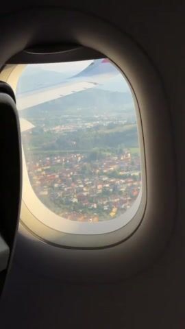 From inside an airplane, a view is captured through the window. The aircraft's wing is partially visible. The outside shows a landscape containing buildings, greenery, and mountains in the distance under a bright sky.