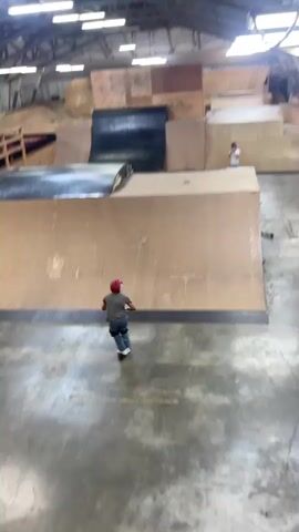 A person on a scooter rides down a ramp in an indoor skatepark. They jump into the air and flip. Another person is standing in the background filming. The skatepark has wooden ramps and walls.