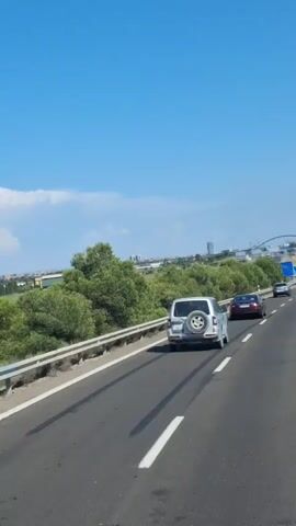 The video shows a view from a moving vehicle on a highway. The road has two lanes in the direction of travel, and there are tire marks visible on the asphalt. The vehicle appears to be driving on the right side of the road. On the left side of the road, there is a concrete barrier and then lush green trees and vegetation. In the distance, there are some buildings and a blue sky with scattered clouds. There are other vehicles on the highway, including a dark-colored car in the foreground and a white SUV in the middle ground. The sun is shining, suggesting it is daytime. The overall impression is a sunny day drive through a scenic area.
