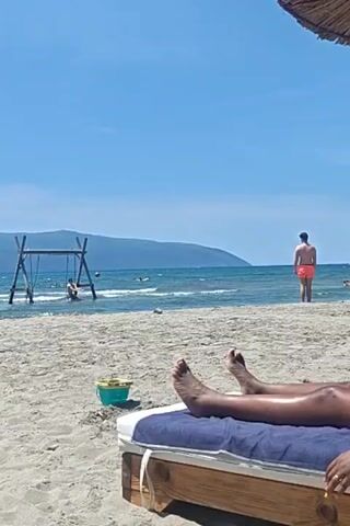 The video shows a beach scene on a sunny day. A person is relaxing on a lounge chair in the foreground. The person is lying down, and only their lower legs and feet are visible. The lounge chair has a wooden frame and a blue and white striped cushion. In the middle ground, there is a wooden swing set standing in the shallow water. A person is on the swing. Further out in the water, there is a person standing in orange swim trunks.  A small green and yellow bucket sits on the sand near the lounge chair. The background features a calm blue sea and a distant, hilly coastline. The sky is clear and blue.