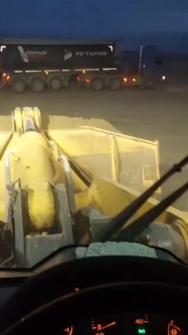 This video shows a first-person perspective from inside a yellow front-end loader. The loader is parked on a dirt or gravel surface, with a pile of yellow material, likely sand or dirt, in front of it. In the background, a large black dump truck with the text "GRÜNWALD" and "РВ-ТАРИФ" is visible. The dump truck has multiple axles and its taillights are illuminated. A blue truck cab is attached to the dump truck's trailer. The sky is dark blue, indicating it is either dawn or dusk. The weather appears to be clear. The scene appears to be a construction site or an industrial area. The time of day is evident from the dark sky and the illuminated vehicle lights. There is no indication of people visible in the footage.