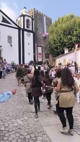 A group of people are walking down a cobblestone street. Some are dressed in historical costumes, appearing to be part of a parade or reenactment. In the background, a church with a white facade and black accents stands next to a stone tower flying flags. Trees and buildings line the street, and a crowd of onlookers watches the procession. The people in historical attire are dressed in tunics, dresses, and head coverings, with some carrying what appear to be drums or musical instruments. The overall atmosphere suggests a historical festival or event taking place in a town square.