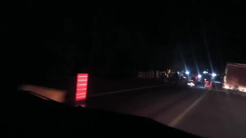 The video was shot at night. The view is from inside a vehicle looking out at a road.  The road is dark, illuminated by the vehicle's headlights. Several road workers are present, wearing high-visibility vests and hard hats. Traffic cones are set up on the road. There's also a barrier present, indicating a lane closure or road work ahead. A large truck is visible on the right side of the road with its lights on. The scene implies road construction or maintenance occurring at night.