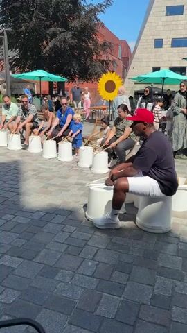 A group of people, including adults and children, are seated in a circle on white buckets, playing drums. They are holding drumsticks and appear to be engaged in a musical activity. Two large sunflower decorations are visible in the background, as well as buildings and trees. The scene takes place outdoors on a paved area.