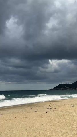The video shows a beach under a cloudy sky. The sand is a light beige color and there are several birds resting on it. The ocean waves are visible, with white foam contrasting against the dark green water. As the video continues, the sky becomes lighter, revealing blue patches. The view then shifts to a concrete platform extending towards the water. The waves crash against the platform, creating splashes.