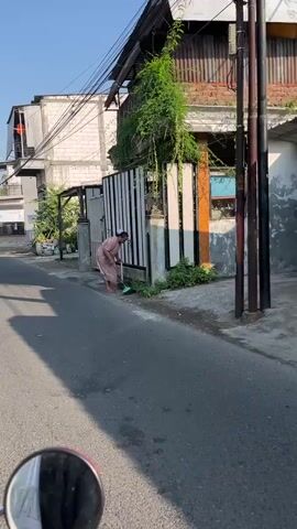 A woman in a pink dress is sweeping the sidewalk in front of a white and black fence. She is using a green broom and a black dustpan. A motorcycle is parked on the street to the left of the scene. The sun is shining and there are shadows on the sidewalk. The background shows a building with a metal roof and some greenery.