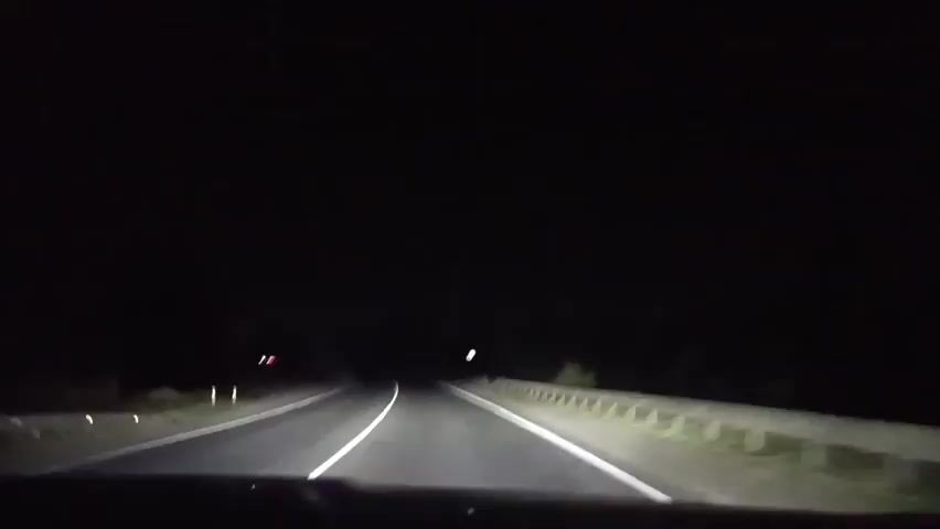A first-person perspective video shows a car driving on a dark, empty road at night. The headlights illuminate the road ahead, revealing two lanes separated by white lines. A guardrail is visible on the right side of the road. The surrounding environment is dark and appears to be rural, with the occasional faint lights or reflective markers visible in the distance. The car continues to move forward at a steady pace.