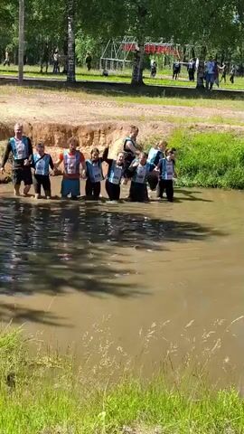 A group of nine people, appearing to be participants in an obstacle course race, stand in a muddy body of water. They are all wearing bib numbers on their vests. The people are holding hands, forming a chain, and raising their free hands in the air, appearing to celebrate or pose for a photo. The water is murky brown and reaches at least to their knees. The background shows a sandy embankment on the left and lush green grass and trees on the right. Further in the background, several other people can be seen, along with some red and white striped structures that resemble an obstacle course. The weather appears to be sunny, with shadows cast on the water. The individuals are diverse in age, with some appearing to be children and others adults. One person on the far right of the group is partially submerged and leaning backward. The overall atmosphere seems to be one of accomplishment and camaraderie.