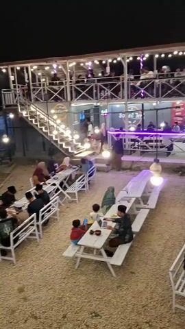 The video shows a busy outdoor dining area at night, illuminated by string lights and a few larger bulbs. There are several white picnic tables and chairs set up on a gravel ground. A two-story building with a balcony and more seating is visible in the background. Many people are seated at the tables, some eating and others conversing. A few motorcycles are parked in the background. The scene appears to be a cafe or restaurant.