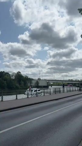 The video shows a peaceful canal scene in Bruges, Belgium. A white motorboat, identified by the text "Le Boat CIRRUS 24" and license plate "NC 8402 S78" on its side, glides through the water. Onboard, two people are visible, one wearing a pink hat and the other a white hat. The boat is leaving a wake as it moves.

On the pedestrian walkway beside the canal, a person is riding a bicycle. The cyclist is wearing a grey t-shirt and sunglasses, and is riding a black bicycle. They are seen from the side as they cycle past.

A concrete bridge spans the canal in the background, with cars visible driving across it. A few streetlights line the walkway, and trees and greenery flank the canal and the far bank. The sky is partly cloudy, with dramatic cumulus clouds interspersed with patches of blue sky, suggesting a dynamic but pleasant day. The overall atmosphere is calm and picturesque, capturing a typical moment of leisure and transport along the historic waterways of Bruges. The time of day appears to be daytime, likely late morning or afternoon, given the brightness and cloud cover.