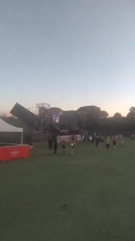 The video pans across an outdoor event space. In the foreground, a grassy field stretches towards a stage structure. Several people are visible milling about on the grass. To the left, a white tent with a red base is set up, displaying the word "ESTRELLA" multiple times. Further back, a large stage is set up with scaffolding, speakers, and illuminated panels. Some of the panels feature graphics and are lit with red and orange lights. A slanted, dark structure is attached to the left side of the main stage. A cell tower stands tall in the background to the left of the stage. The sky is clear and appears to be dusk or early evening. The overall impression is of an outdoor concert or festival setting before or during the early stages of the event.
