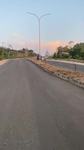 A low-angle, first-person perspective video captured on a paved road. The road curves to the left and is bordered by a concrete curb with dry vegetation. Tall, modern streetlights with two arms are spaced along the right side of the road. In the distance, a small truck with two people in the cab and one standing in the back drives away from the camera on the same road. The sky is a light blue with a few wispy pink and orange clouds, suggesting either sunrise or sunset. The environment appears somewhat undeveloped, with some trees and hilly terrain visible in the background.